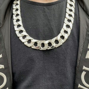 May include: A silver chain necklace with a large, chunky link design. The chain is worn over a black t-shirt.