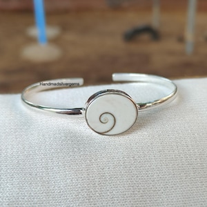 May include: A silver cuff bracelet with a white oval centerpiece. The centerpiece features a spiral design in a darker color. The bracelet is displayed on a white surface. The text "Handmadsilvergems" is visible on the bracelet.