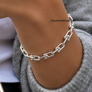 Könnte beinhalten: Ein silbernes Armband mit einem Kettenglied-Design. Das Armband besteht aus glänzendem Metall und wird am Handgelenk einer Person getragen. Das Armband hat eine Reihe von U-förmigen Gliedern, die durch kleine, runde Perlen verbunden sind. Der Text "Handmadsilvergems" ist sichtbar.