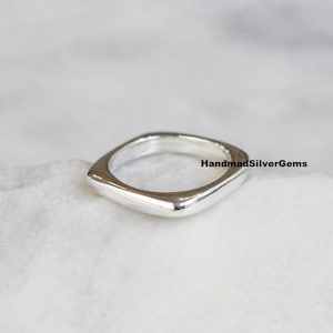 Puede incluir: Un anillo de plata pulida con forma cuadrada. El anillo tiene bordes redondeados y una superficie lisa y reflectante. El texto "HandmadSilverGems" es visible en el lado derecho de la imagen. El anillo está sobre un fondo claro.