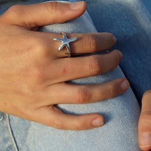 May include: A silver starfish ring. The ring features a detailed starfish design. The ring is worn on a finger. The background is a person's blue jeans.