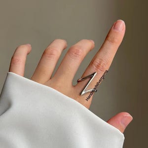 May include: A silver-colored ring with a geometric Z-shaped design. The ring is worn on a finger, with the design wrapping around the finger. The ring is shiny and reflects light. The text "Handmadsilvergems" is visible on the ring.
