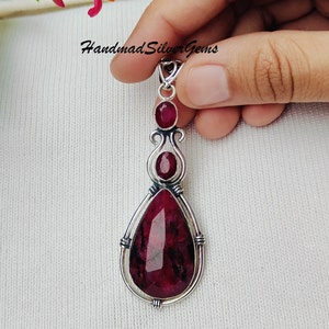 以下が含まれることがあります： 3つのルビーの宝石が付いた手作りのシルバーペンダント。ペンダントは、下部に大きな洋梨型のルビー、2つの小さな楕円形のルビー、装飾的なシルバーフィリグリーが特徴です。「Handmad Silver Gems」というテキストが表示されます。