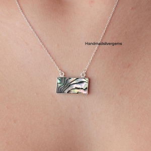 Op de afbeelding: Een zilveren ketting met een rechthoekige hanger. De hanger heeft een kleurrijk abalone schelpontwerp met tinten groen, blauw en roze. De ketting wordt op de borst van een persoon getoond.