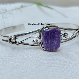 Pode incluir: Uma pulseira de punho prateada com uma pedra preciosa roxa retangular no centro. A pulseira apresenta redemoinhos decorativos e o texto "HandmadSilverGems". A pedra preciosa tem uma aparência marmorizada.