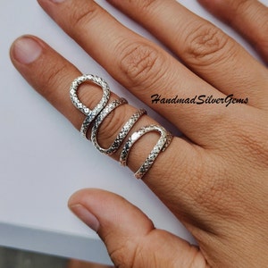May include: A silver-colored ring wraps around a finger in a snake-like design. The ring has a textured surface and is coiled to create a unique, eye-catching accessory. The text "HandmadSilverGems" is visible.