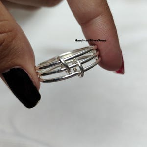 Peut inclure: Une bague en argent au design unique. La bague présente trois bandes parallèles reliées par deux barres verticales. La bague est tenue entre les doigts sur fond blanc. Le texte "HandmadSilverGems" est visible.