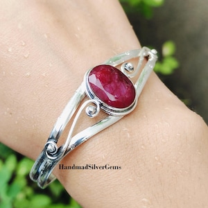 Może przedstawiać: Srebrna bransoletka z owalnym, głębokim, czerwonym kamieniem szlachetnym. Bransoletka ma otwartą konstrukcję z dekoracyjnymi wirami i napisem "HandmadSilverGems" na opasce.