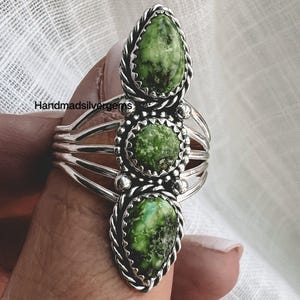May include: A silver ring with three green stone settings. The stones are teardrop and oval shaped, with a textured appearance. The ring has a braided border around each stone and multiple bands on the ring's base. The text "Handmadesilvergems" is visible.