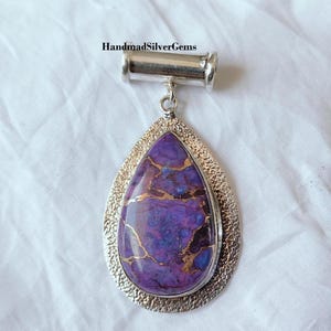 May include: A teardrop-shaped pendant featuring a striking purple stone with gold veining, set within a textured silver frame. The pendant is suspended from a silver bail. The text "HandmadSilverGems" is visible.
