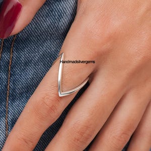 May include: A silver V-shaped ring is worn on a finger. The ring has a minimalist design, with a smooth, polished surface. The text "Handmadsilvergems" is visible in the background. The ring is a piece of jewelry.