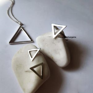 May include: A silver geometric jewellery set featuring a triangle pendant necklace, a ring, and a pair of stud earrings. The jewellery is displayed on white stones against a white background. The ring and earrings have a hollow triangle design.