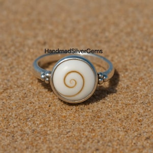Puede incluir: Anillo de plata con una pieza central circular blanca con un diseño en espiral en tonos marrones. El anillo está sobre un fondo arenoso, con el texto "HandmadSilverGems" visible.