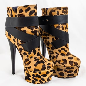 May include: A pair of high-heeled ankle boots with a leopard print pattern. The boots feature a platform sole, a black strap across the ankle, and a stiletto heel. The boots are approximately 6 inches tall.
