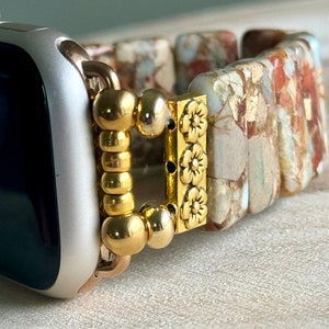 May include: A gold-toned watch band with a floral design and a beaded clasp. The band is made of a light brown material with a marbled pattern.