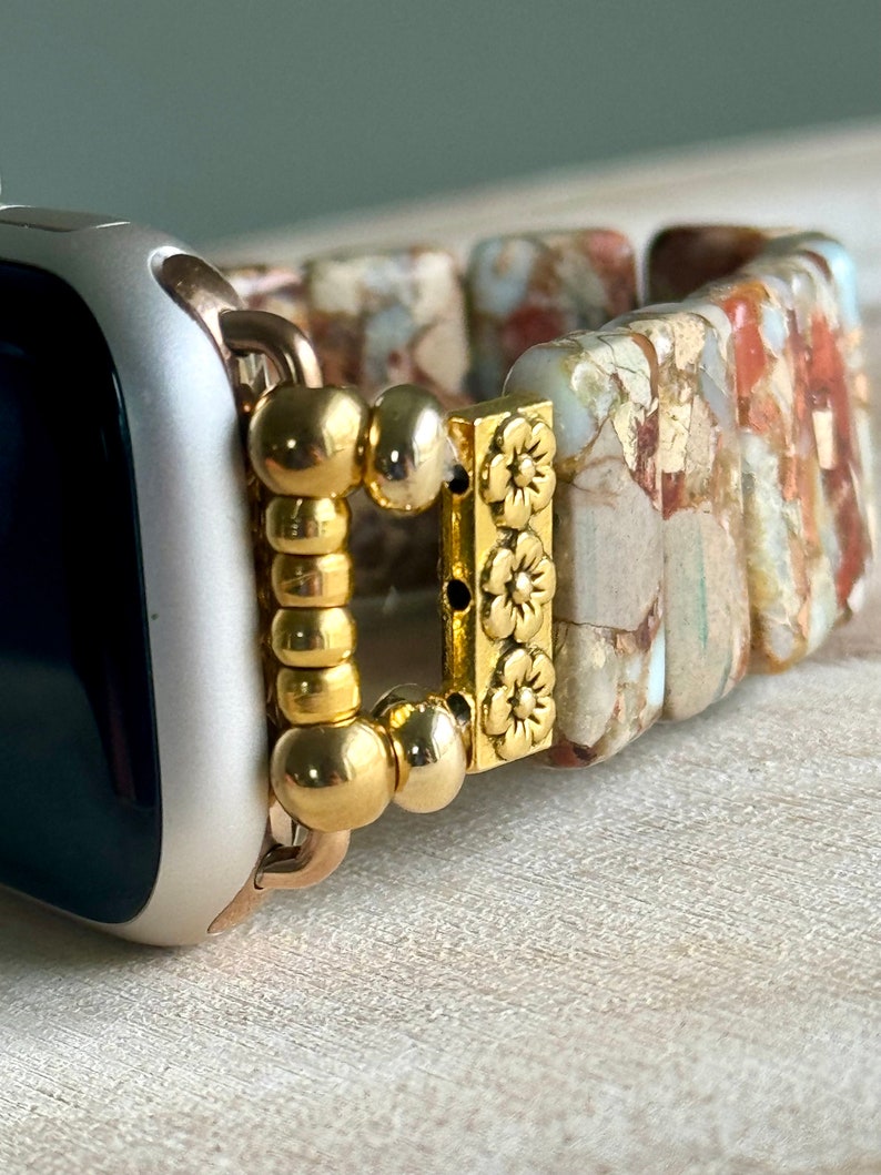 May include: A gold-toned watch band with a floral design and a beaded clasp. The band is made of a light brown material with a marbled pattern.