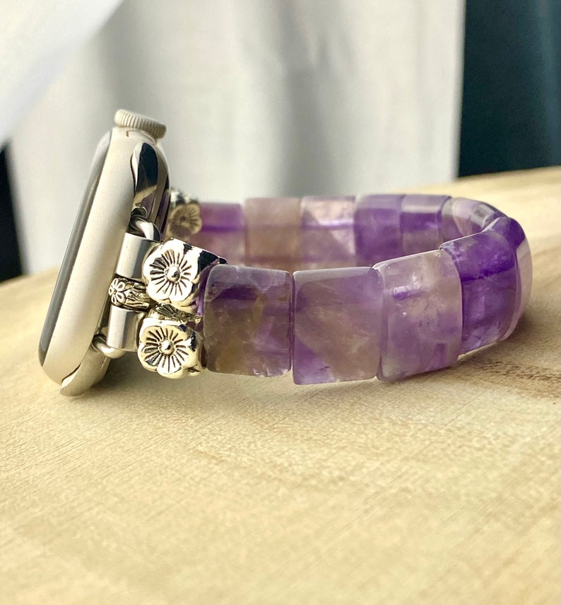 May include: A silver watch with a purple amethyst gemstone band. The band is made of square-shaped beads and has silver flower accents.