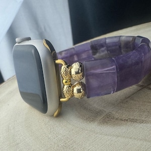 May include: A purple amethyst beaded watch band with gold accents. The band is made of square-shaped beads and has a gold clasp.