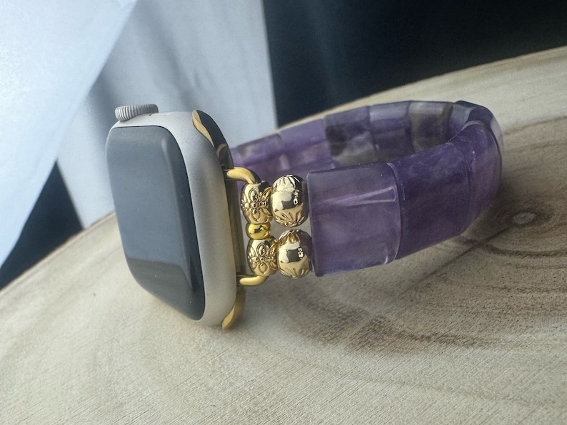 May include: A purple amethyst beaded watch band with gold accents. The band is made of square-shaped beads and has a gold clasp.