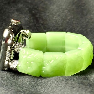 Pode incluir: Uma pulseira verde claro com fecho prateado. A pulseira é feita de contas quadradas translúcidas. O fecho tem desenhos florais e geométricos. A pulseira está sobre uma superfície cinza escuro.