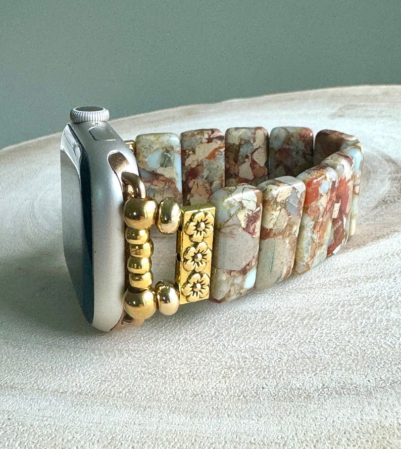 May include: A beige and brown patterned Apple Watch band with a gold clasp and decorative beads. The band is made of stone and has a floral design.