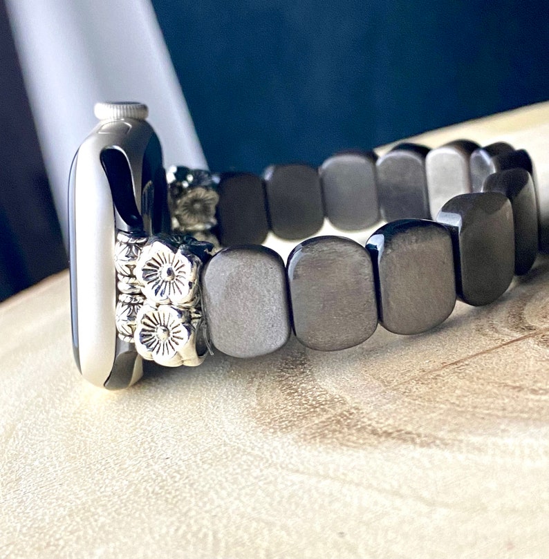 May include: A black and silver Apple Watch band made of square-shaped beads. The band has a silver floral design on the watch face side.