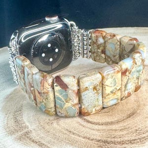 May include: A silver and beige Apple Watch band made of rectangular, patterned stones. The band is attached to a silver watch with a black face.