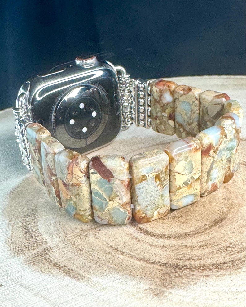 May include: A silver and beige Apple Watch band made of rectangular, patterned stones. The band is attached to a silver watch with a black face.