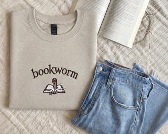 May include: A beige crewneck sweatshirt featuring the embroidered text bookworm in dark brown letters above a small graphic of a brown worm wearing glasses inside an open book, paired with light blue denim jeans and an open hardcover book.