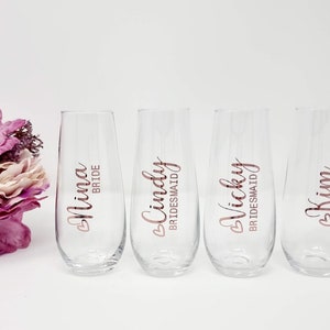 May include: Four clear stemless champagne flutes with rose gold lettering. The glasses are labeled "Nina Bride", "Cindy Bridesmaid", "Vicky Bridesmaid", and "Kim Maid of Honour". A bouquet of pink and purple flowers is on the left.