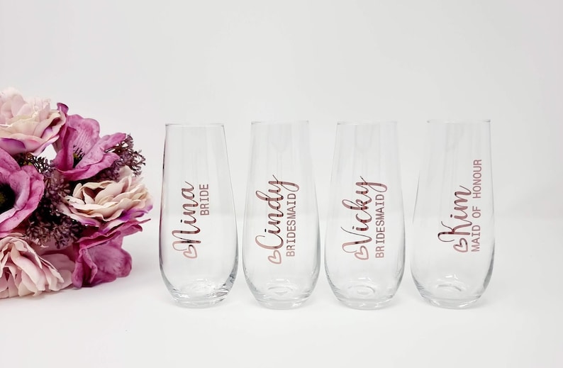 May include: Four clear stemless champagne flutes with rose gold lettering. The glasses are labeled "Nina Bride", "Cindy Bridesmaid", "Vicky Bridesmaid", and "Kim Maid of Honour". A bouquet of pink and purple flowers is on the left.