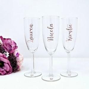May include: Three clear champagne flutes with names Lauren, Nicola, and Kirstie in a copper-colored script. Each glass has a heart etched on the base. A bouquet of purple flowers is in the background.