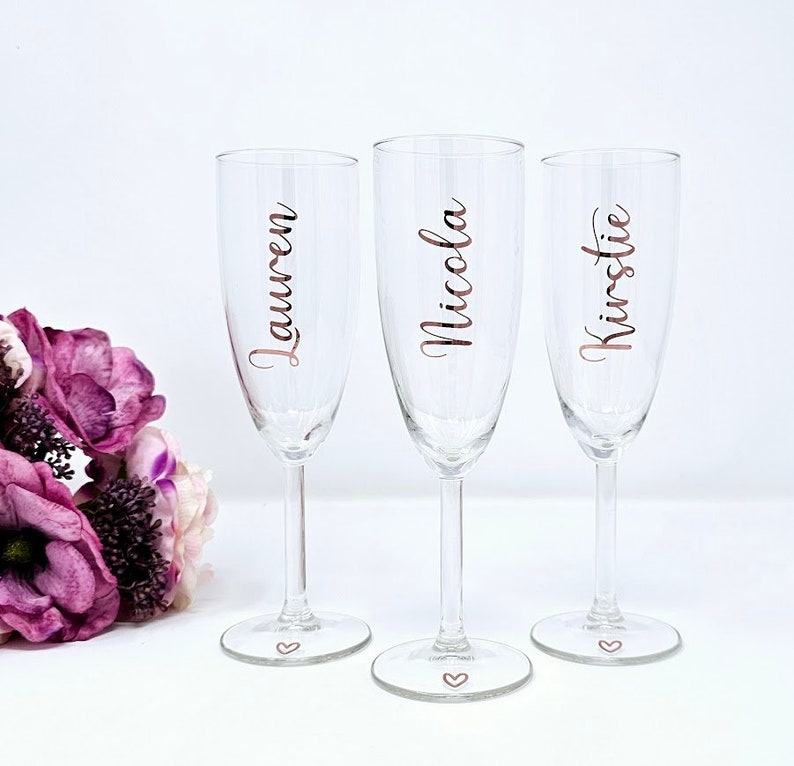 May include: Three clear champagne flutes with names Lauren, Nicola, and Kirstie in a copper-colored script. Each glass has a heart etched on the base. A bouquet of purple flowers is in the background.