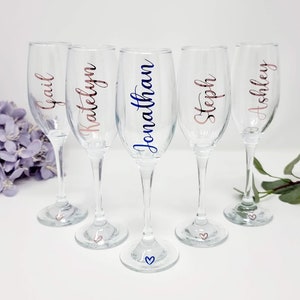 May include: Five clear champagne flutes with personalized names in various colors. The names Gail, Katelyn, Jonathan, Steph, and Ashley are displayed in rose gold and blue script. Each flute has a small heart design on the base.