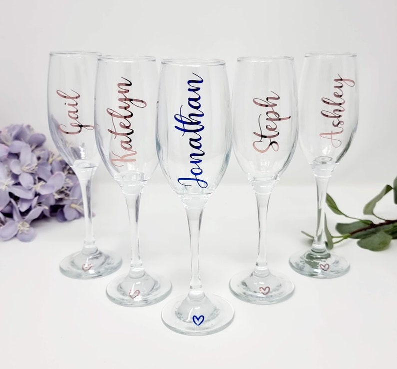 May include: Five clear champagne flutes with personalized names in various colors. The names Gail, Katelyn, Jonathan, Steph, and Ashley are displayed in rose gold and blue script. Each flute has a small heart design on the base.