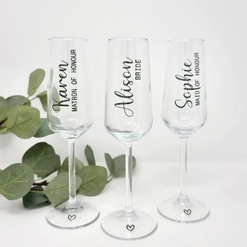 May include: Three clear champagne flutes with black text. The flutes are labeled "Karen MATRON OF HONOUR", "Alison BRIDE", and "Sophie MAID OF HONOUR". Each flute has a small heart etched on the base. Green foliage is in the background.