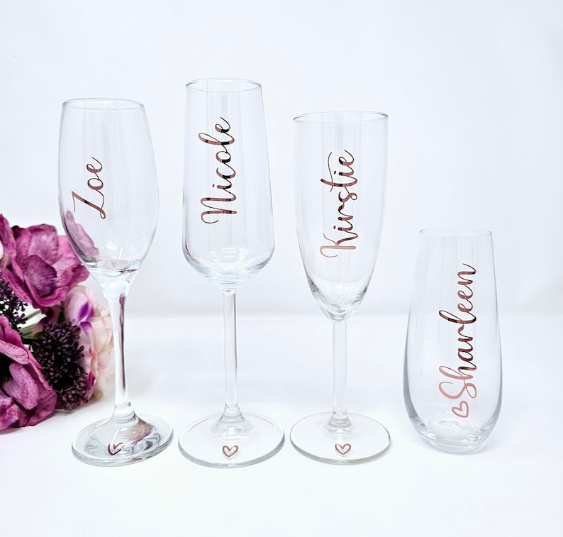 May include: Four clear champagne flutes with names in rose gold script: Zoe, Nicole, Kirstie, and Sharleen. Each glass has a small heart at the base. A bouquet of purple flowers is in the background.