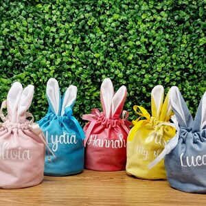 May include: Five pastel-coloured bunny-shaped drawstring bags with embroidered names: Olivia, Lydia, Hannah, My, and Lucas. The bags are arranged in a row on a wooden surface in front of a green leafy backdrop.