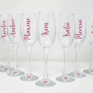 May include: A set of eight clear champagne flutes with names in pink script. Each glass has a heart at the base. Names include Bernie, Kirsten, Amber, Maxine, Kim, Katie, Mercia, Cat, and Bryony. Ideal for a bridal party or special event.