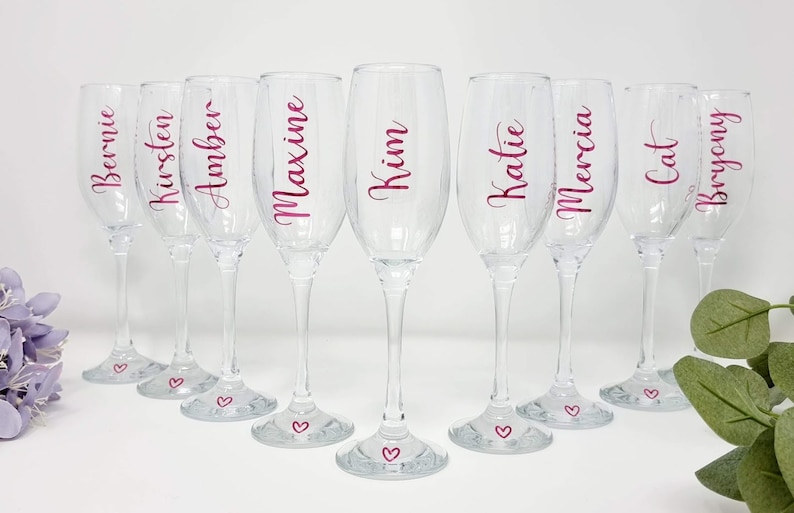 May include: A set of eight clear champagne flutes with names in pink script. Each glass has a heart at the base. Names include Bernie, Kirsten, Amber, Maxine, Kim, Katie, Mercia, Cat, and Bryony. Ideal for a bridal party or special event.