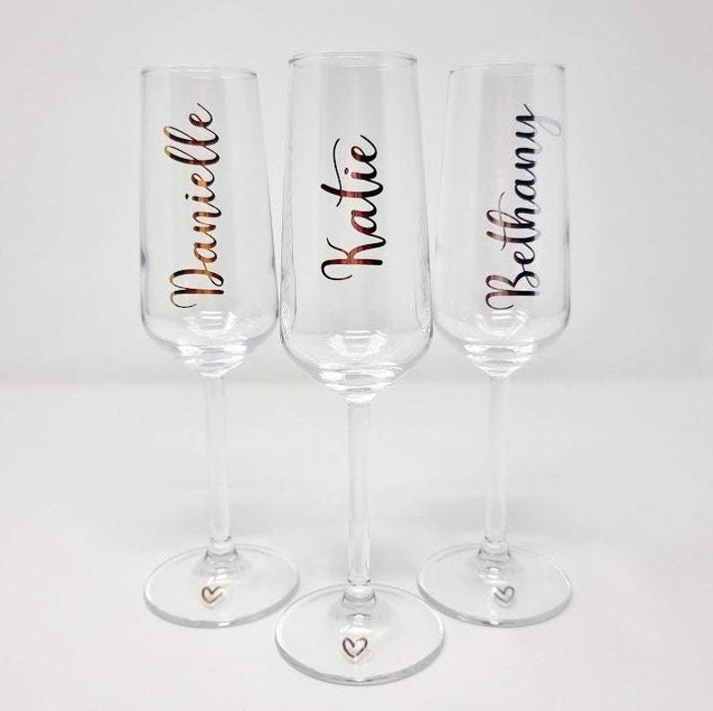 May include: Three clear champagne flutes with names Danielle, Katie, and Bethany in a copper-colored script. Each flute has a small heart at the base. The glasses are on a white surface.