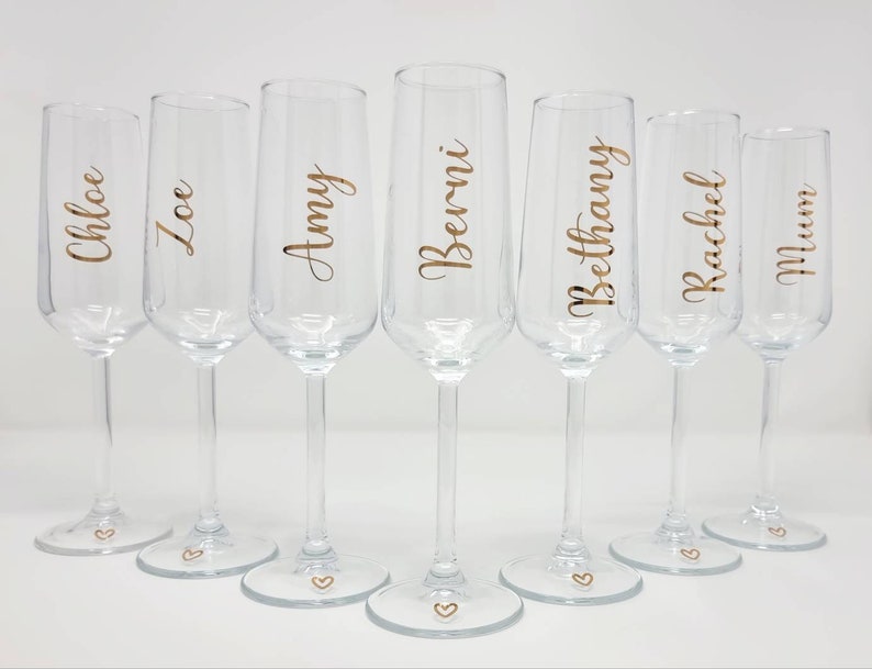 May include: Seven clear champagne flutes with gold lettering. Each glass has a different name: Chloe, Zoe, Amy, Berni, Bethany, Rachel, and Mum. A small gold heart is at the base of each glass.