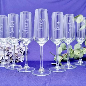 May include: Seven clear champagne flutes with names etched on them: Kate, Sally, Henry, Lucy, Ben, Laneta, and Rob. The flutes are arranged on a purple surface, with a floral arrangement and greenery in the background. Each flute has a heart design on the base.