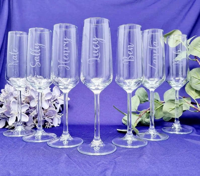 May include: Seven clear champagne flutes with names etched on them: Kate, Sally, Henry, Lucy, Ben, Laneta, and Rob. The flutes are arranged on a purple surface, with a floral arrangement and greenery in the background. Each flute has a heart design on the base.