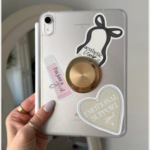 May include: A white iPad case with a variety of stickers. The stickers include a pink tube of lip gloss with the text "I'm just a girl", a black and white bow with the text "aesthetic club", a gold-colored phone ring, and a beige heart with the text "Emotional Support Donut".