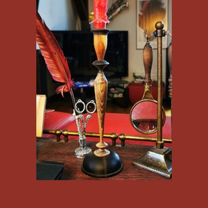 May include: A wooden candle holder with a black base and a red candle. The candle holder is on a wooden table with a feather quill, scissors, and a magnifying glass.