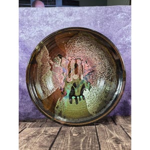 May include: A handmade ceramic bowl with a unique glaze. The bowl features a dark brown rim and a center with pink, green, and white speckled patterns. The bowl is round and sits on a wooden surface.