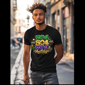 Puede incluir: Camiseta negra con un diseño gráfico colorido que presenta el número "504" en verde, amarillo y morado, junto con las palabras "New Orleans". El diseño incluye elementos temáticos de Mardi Gras.