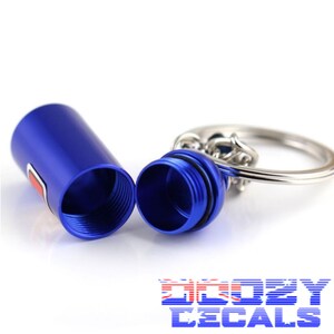May include: A blue metal keychain pill container with a screw-on lid. The container is open and the lid is off. The keychain is attached to a silver metal ring.