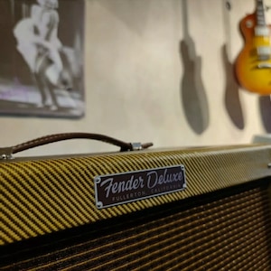 Puede incluir: Un amplificador de guitarra Fender Deluxe de estilo vintage con una cubierta de tolex marrón y una rejilla de tela dorada. El amplificador tiene una manija de cuero marrón y una placa de identificación plateada que dice "Fender Deluxe Fullerton, California".